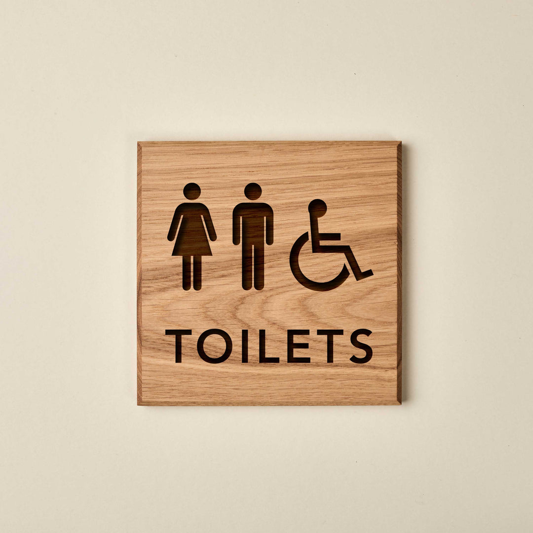 Square oak all-gender and accessible restroom sign. It features engraved female, male, and wheelchair icons positioned above the word "TOILETS." The sign has a warm, natural wood finish with a prominent grain.