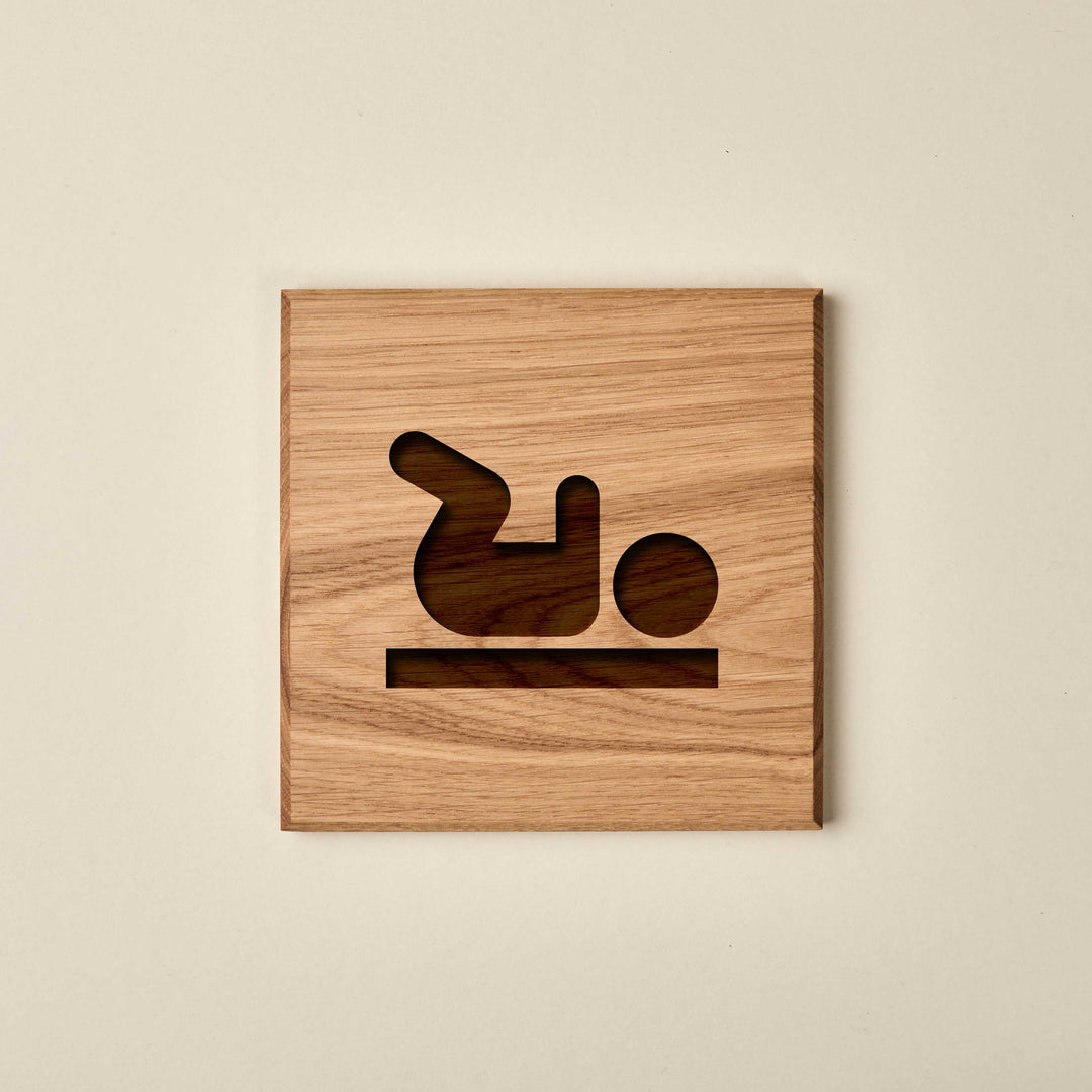 Square oak baby changing station sign. Features an engraved infant silhouette and changing table icon with a dark recessed wood finish.