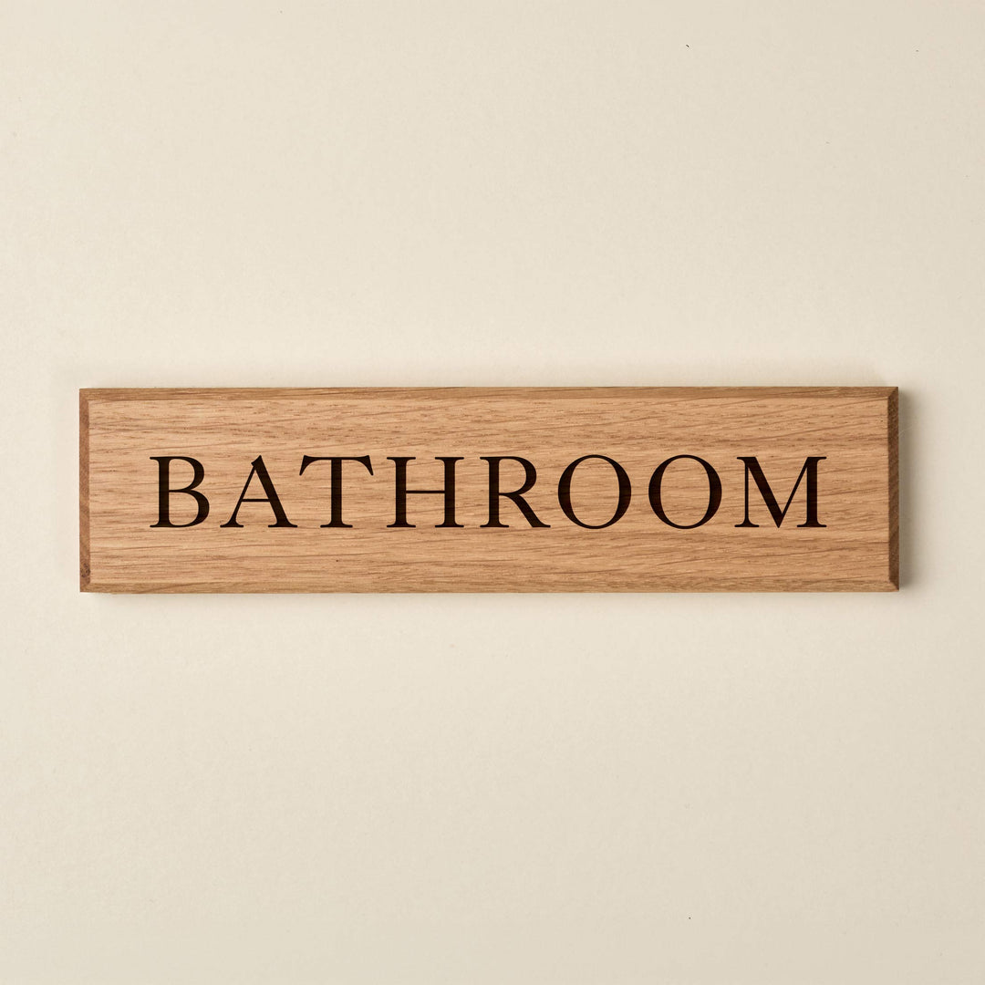 A rectangular oak wood sign featuring a natural horizontal grain and clean, bevelled edges. The word "Bathroom" is engraved in a classic serif font. The recessed lettering reveals a darker wood grain beneath, creating a sophisticated stencil effect against the light oak surface.