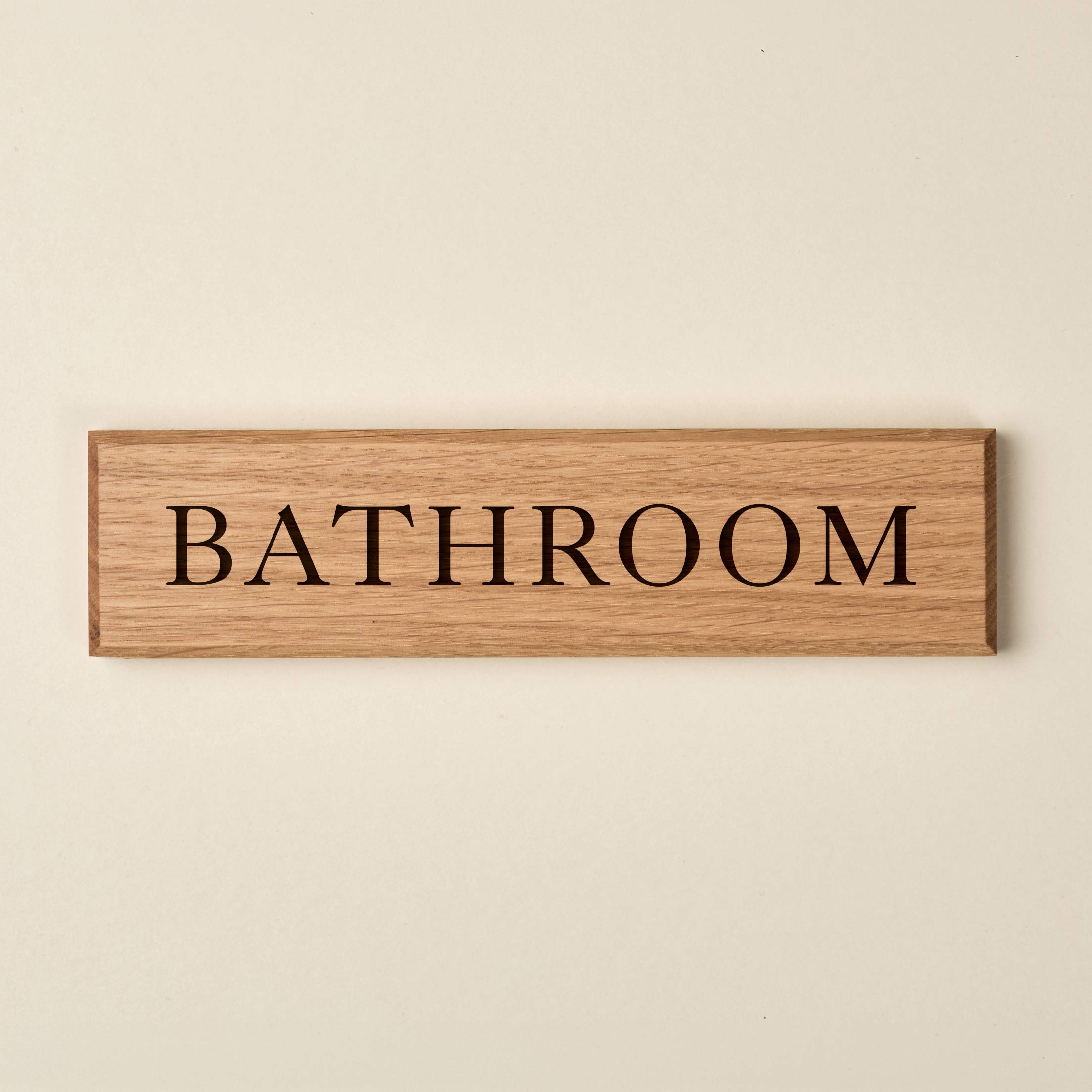A rectangular oak wood sign featuring a natural horizontal grain and clean, bevelled edges. The word "Bathroom" is engraved in a classic serif font. The recessed lettering reveals a darker wood grain beneath, creating a sophisticated stencil effect against the light oak surface.