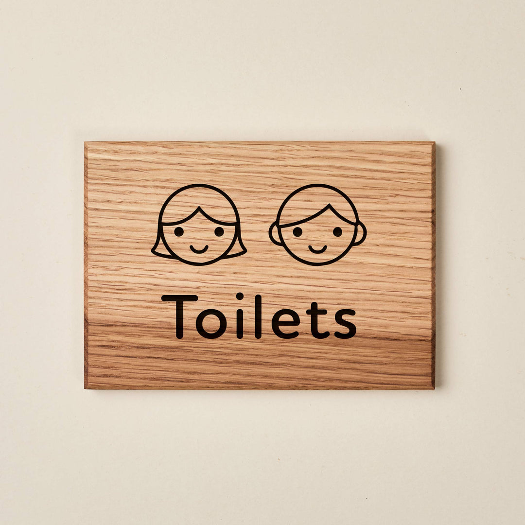 Rectangular oak children's restroom sign. Features simple engraved line drawings of smiling boy and girl faces above the word "Toilets" in a rounded font. The natural wood finish has a visible grain and dark engraved detailing.
