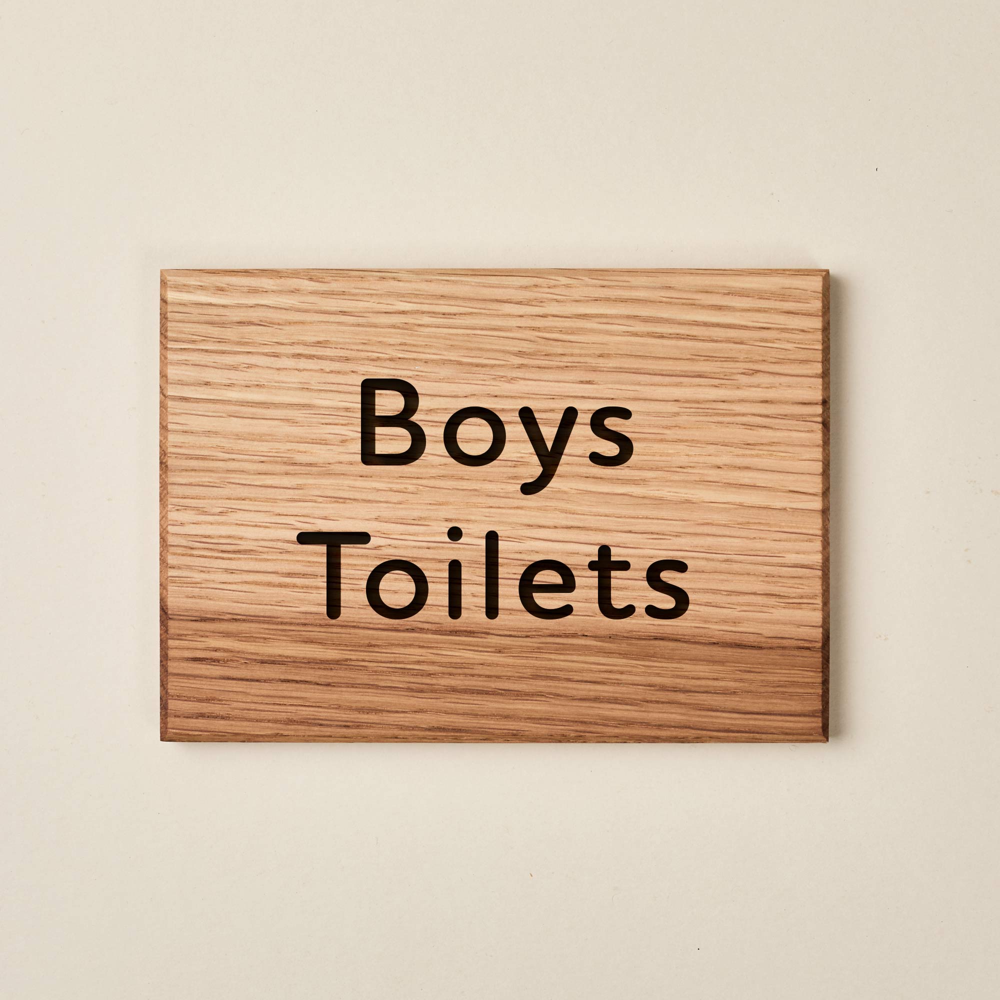 A rectangular oak wood sign with a natural horizontal grain. The words "Boys Toilets" are engraved in a friendly, rounded sans-serif font across two lines. The dark, recessed lettering provides a clear contrast against the light-colored wood surface, creating a welcoming and legible sign for a school or nursery setting.