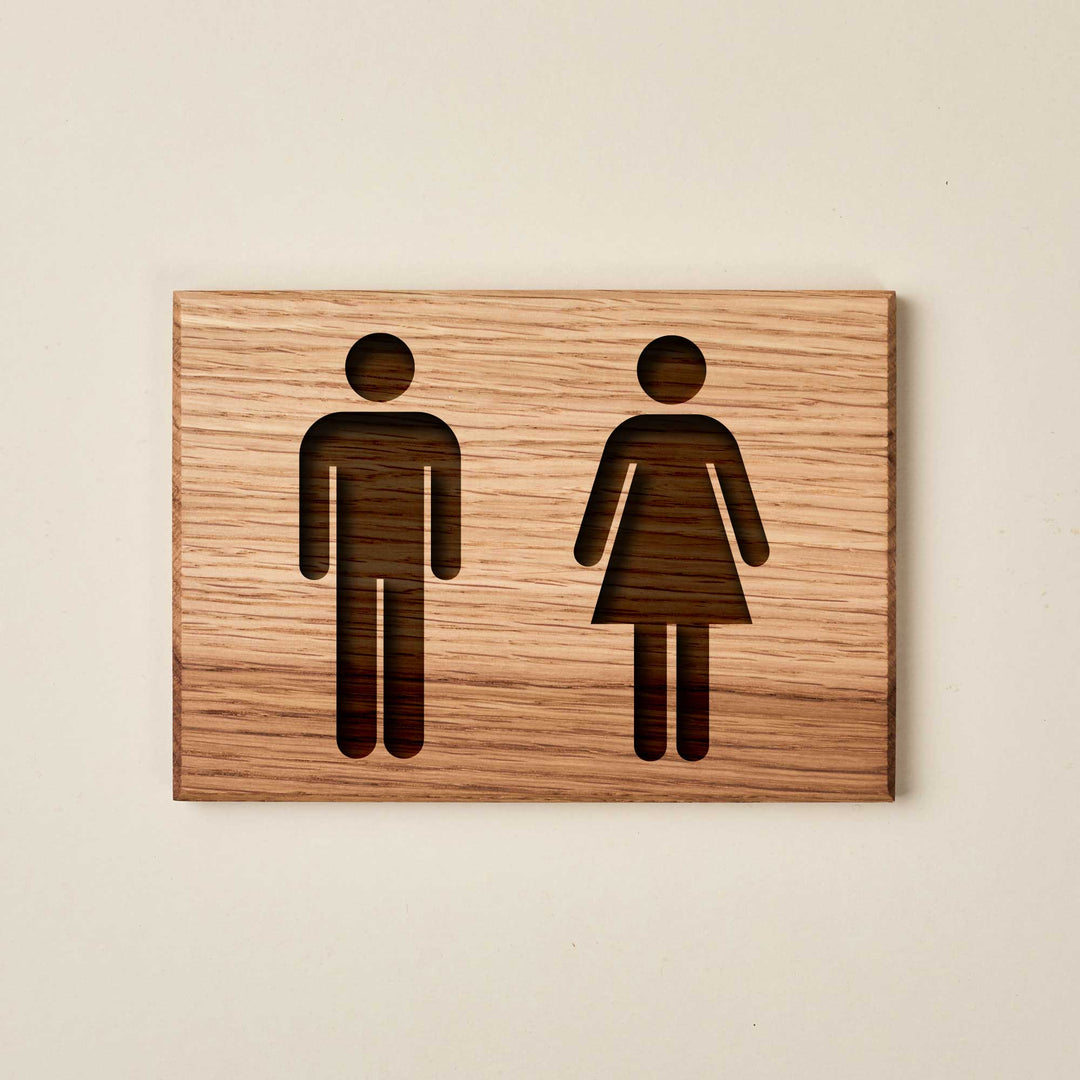 Minimalist oak wood restroom sign featuring engraved male and female symbols. The natural wood grain runs horizontally across the plaque.