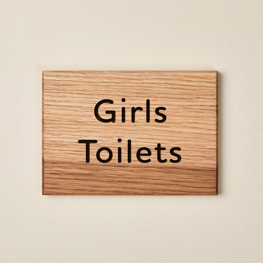 A rectangular oak wood sign with a natural horizontal grain. The words "Girls Toilets" are engraved in a friendly, rounded sans-serif font across two lines. The dark, recessed lettering provides a clear contrast against the light-colored wood surface, creating a welcoming and legible sign for a school or nursery setting.