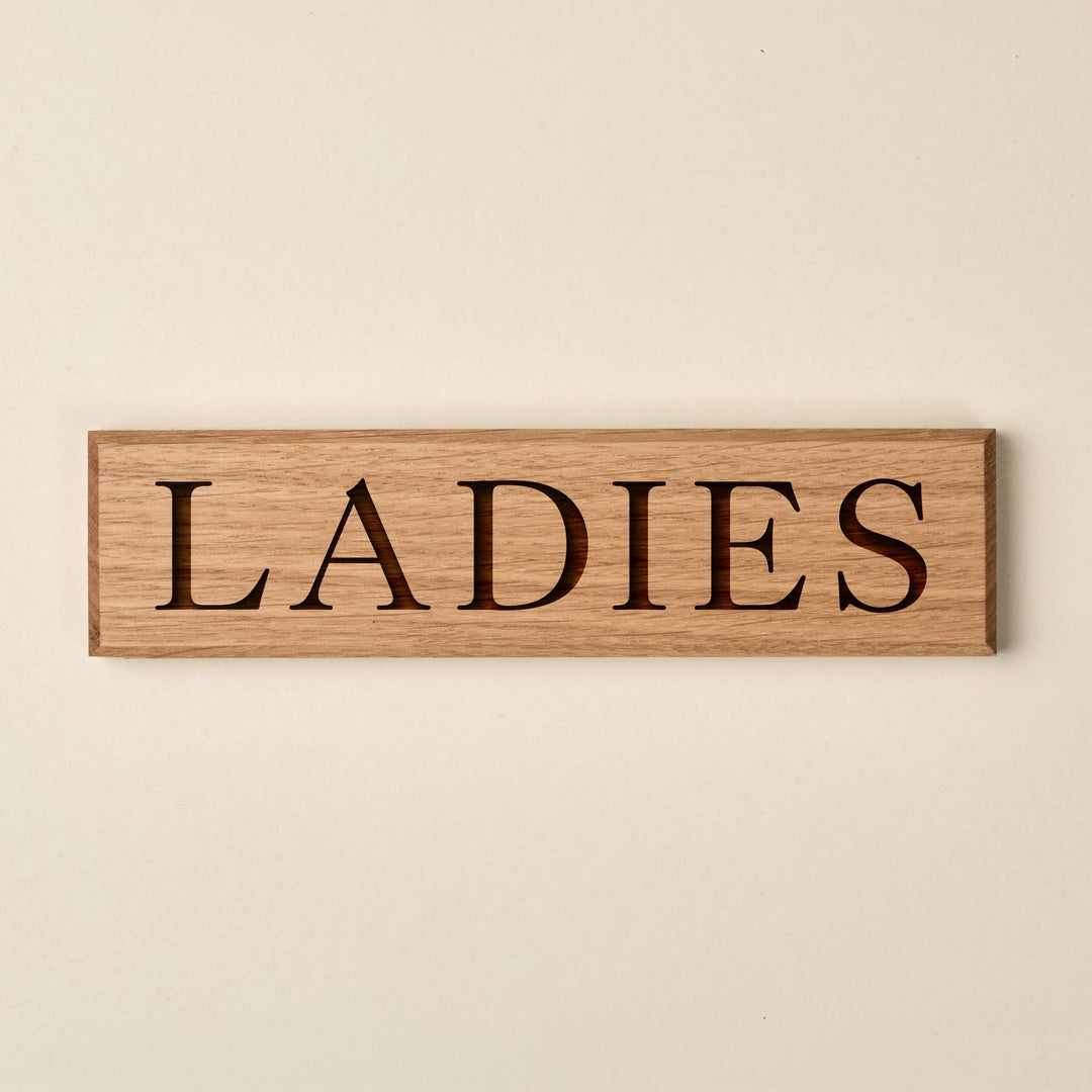 Horizontal oak "LADIES" door sign featuring engraved serif lettering. The sign showcases a warm, natural wood finish with a visible grain and dark recessed text.