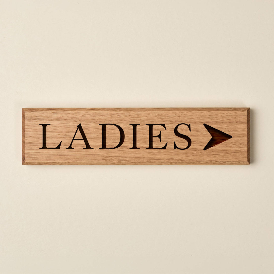 Slimline oak "LADIES" directional sign featuring engraved serif lettering and a right-pointing arrow. The natural wood finish has a visible horizontal grain and dark recessed text.
