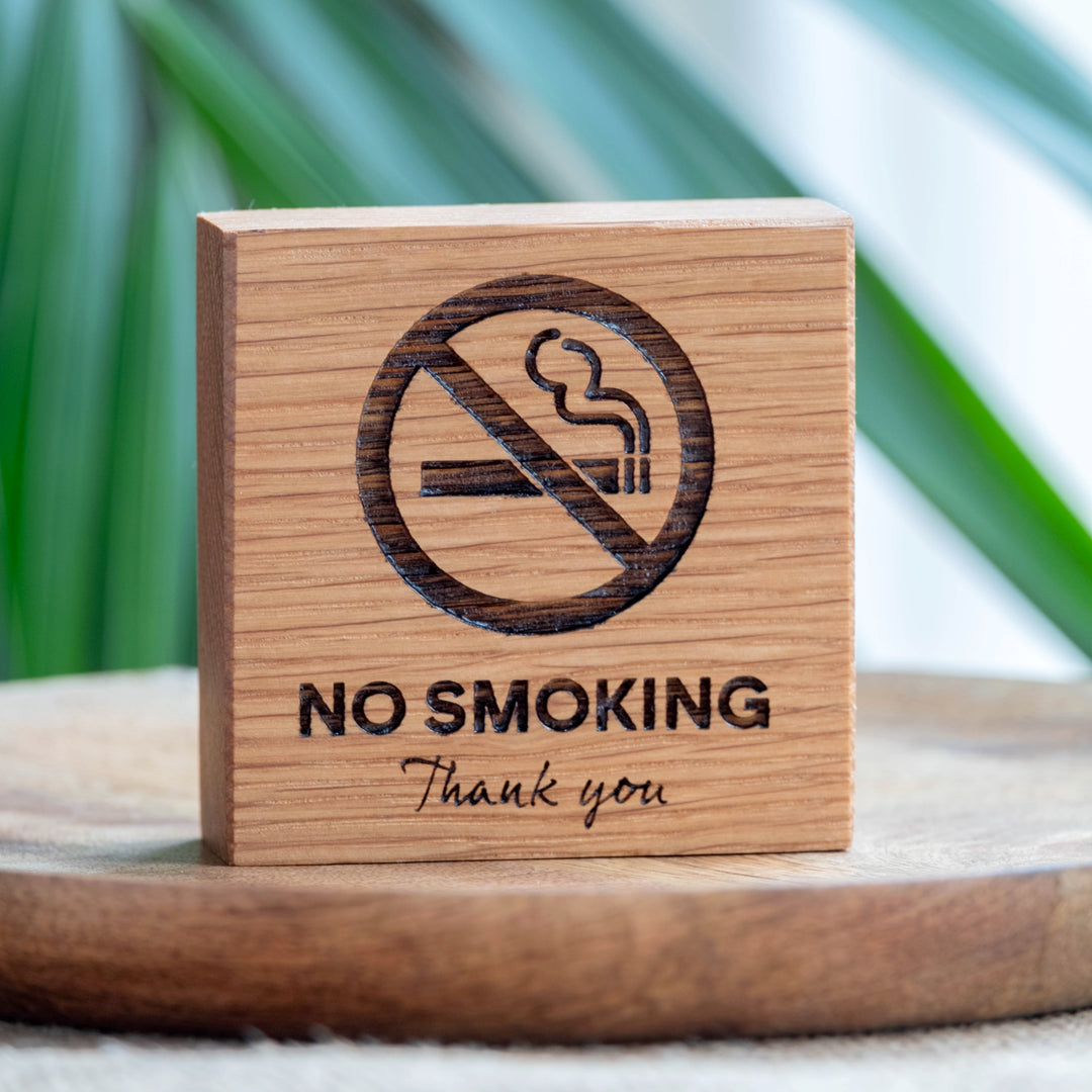 A wooden no smoking sign with the symbol and text 'NO SMOKING Thank you' engraved on it, displayed on a wooden surface with a plant in the background.