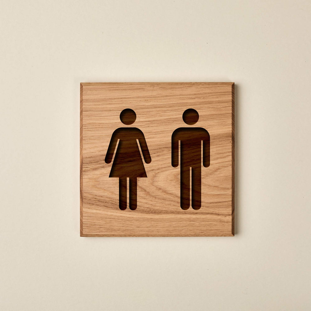 A squares wooden sign made of light-colored oak with a visible horizontal grain. Two gender symbols—a male figure on the left and a female figure on the right—are engraved revealing a darker wood layer or shadow underneath.
