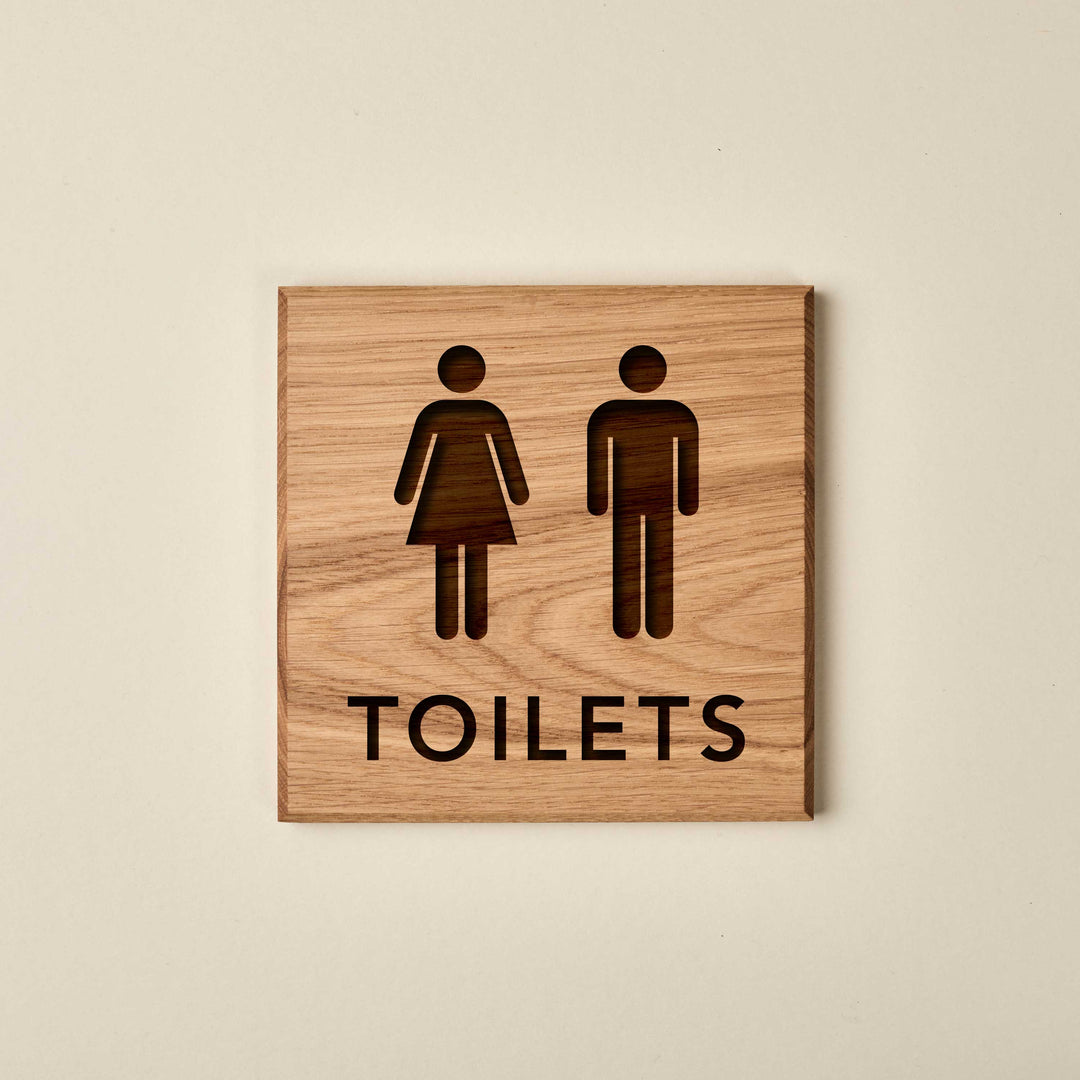 Square oak restroom sign featuring engraved male and female icons above the word "TOILETS." The sign is made of light-colored wood with a visible grain and dark recessed lettering.