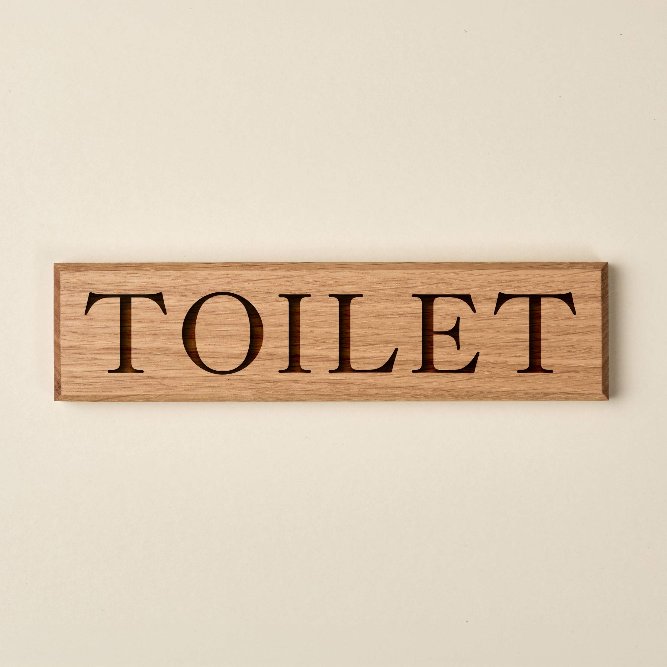 A long, narrow rectangular oak wood sign featuring a natural horizontal grain and clean, beveled edges. The word "TOILET" is engraved in a classic, high-contrast serif font. The recessed lettering reveals a darker wood grain beneath, creating a sophisticated stencil effect against the light oak surface.