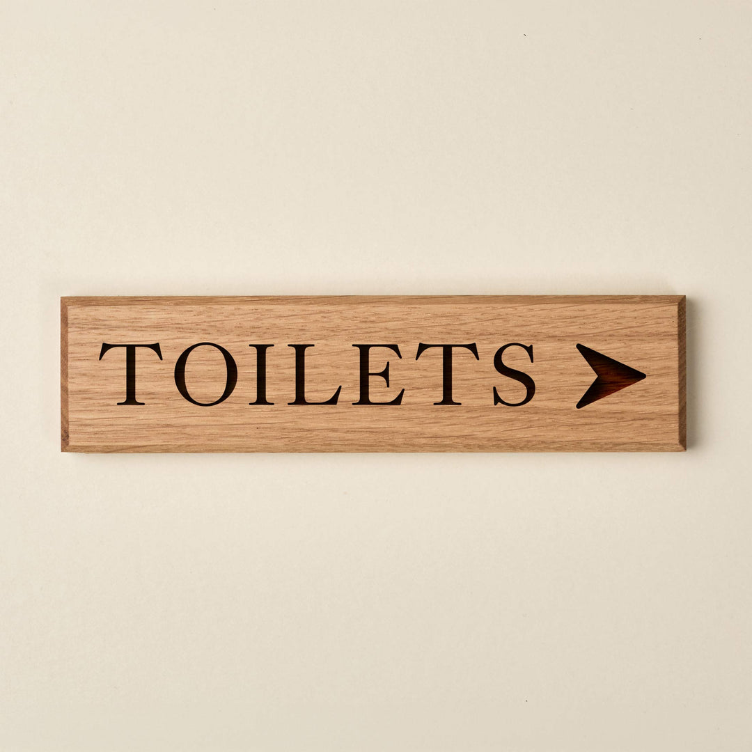 Slimline oak "TOILETS" directional sign featuring engraved serif lettering and a right-pointing arrow. The natural wood finish has a visible horizontal grain and dark recessed text.
