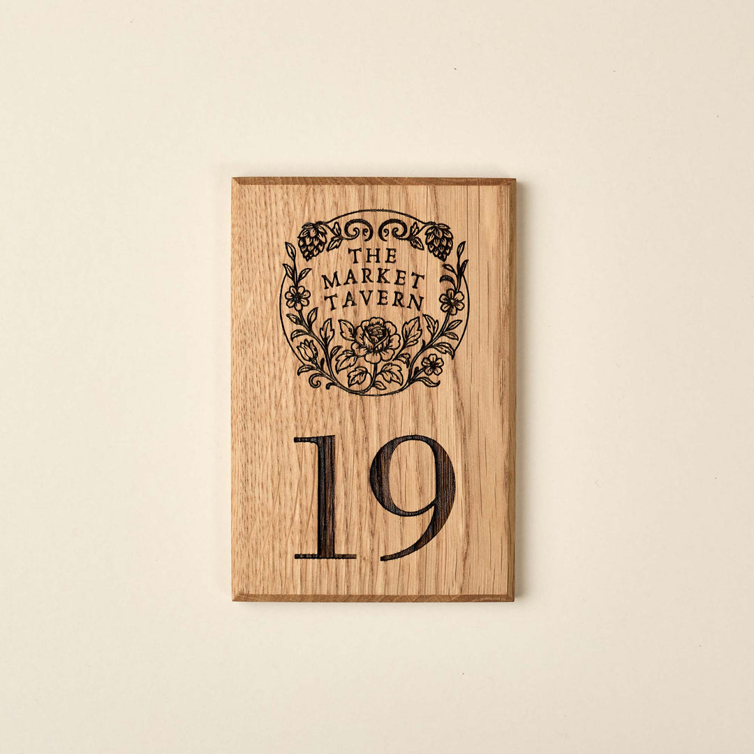 A rectangular solid oak hotel room door sign, engraved with the number 19 and a detailed floral brand logo for The Market Tavern